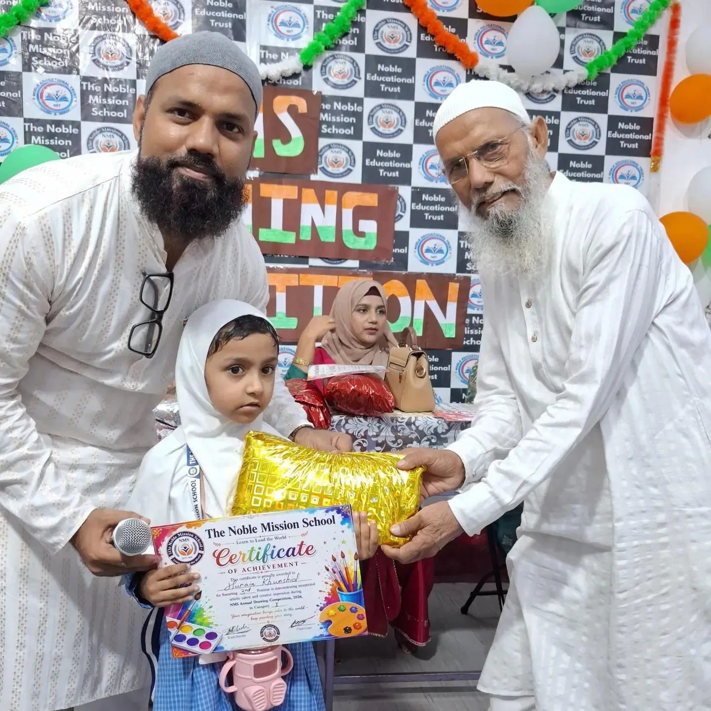 A young student in uniform holding a certificate of achievement while two teachers present her prize in front of the NMS Drawing Competition backdrop